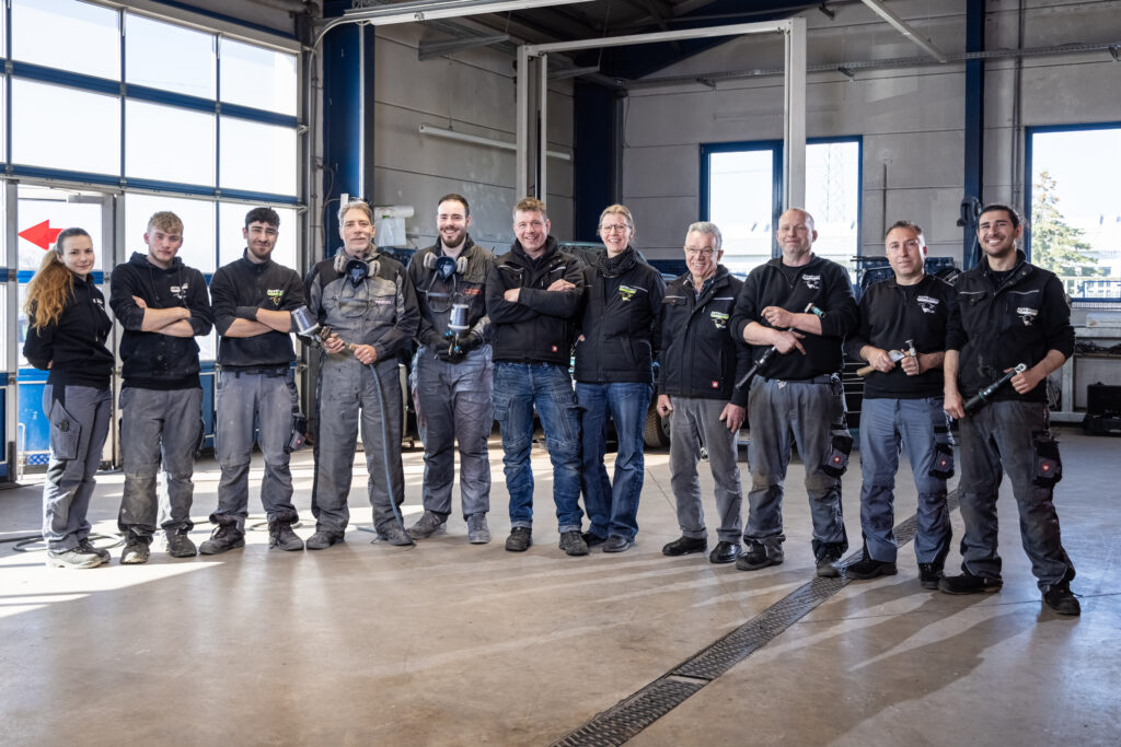 Das Bild zeigt eine Gruppe von Mitarbeitenden in Arbeitskleidung, die in einer Werkstatthalle nebeneinander stehen. Einige halten Werkzeuge oder Spritzpistolen in den Händen.